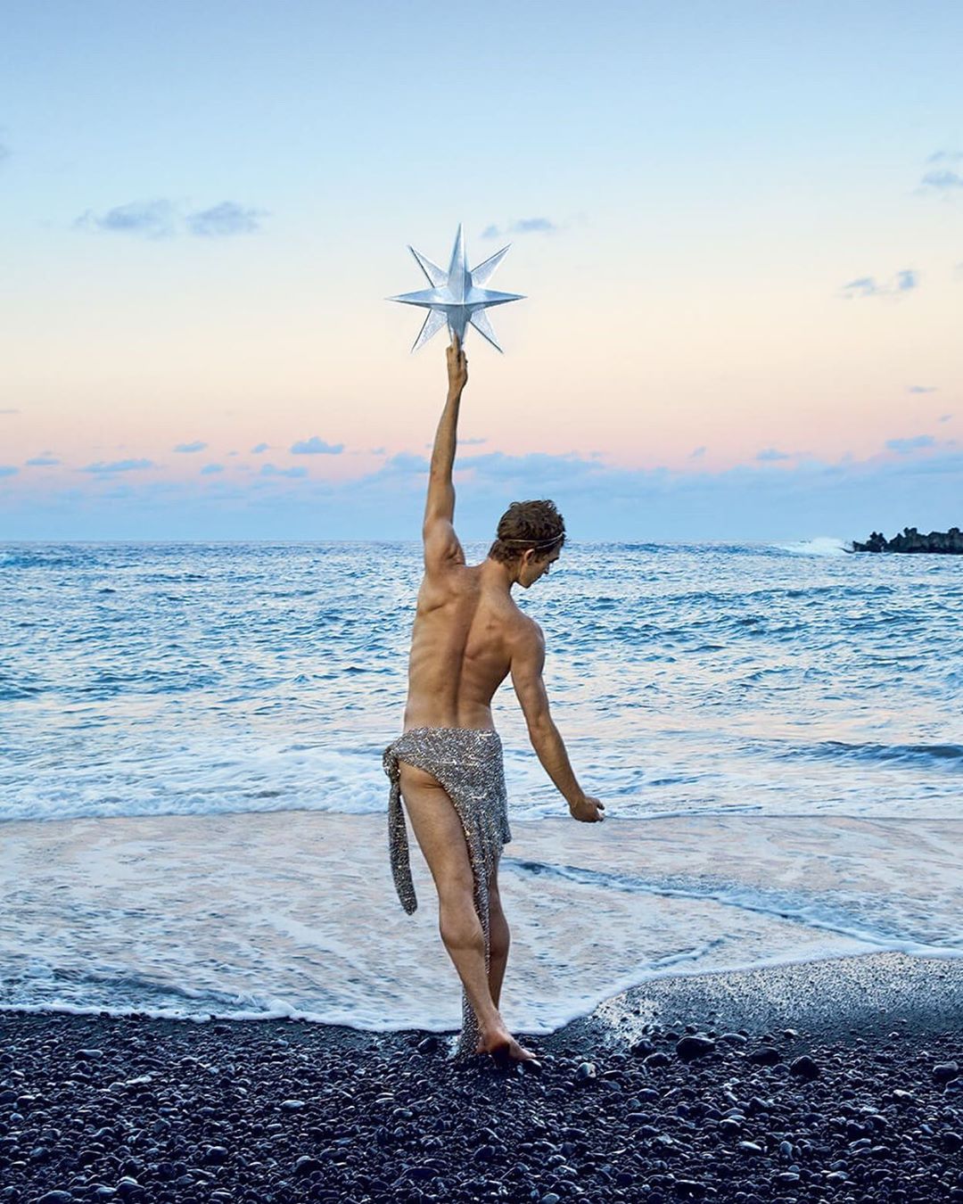 Adult male stands on black sand beach, holding star prop. He faces away from the camera, ocean and sky in background with sunset colors.