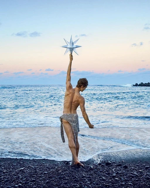 Adult male stands on black sand beach, holding star prop. He faces away from the camera, ocean and sky in background with sunset colors.