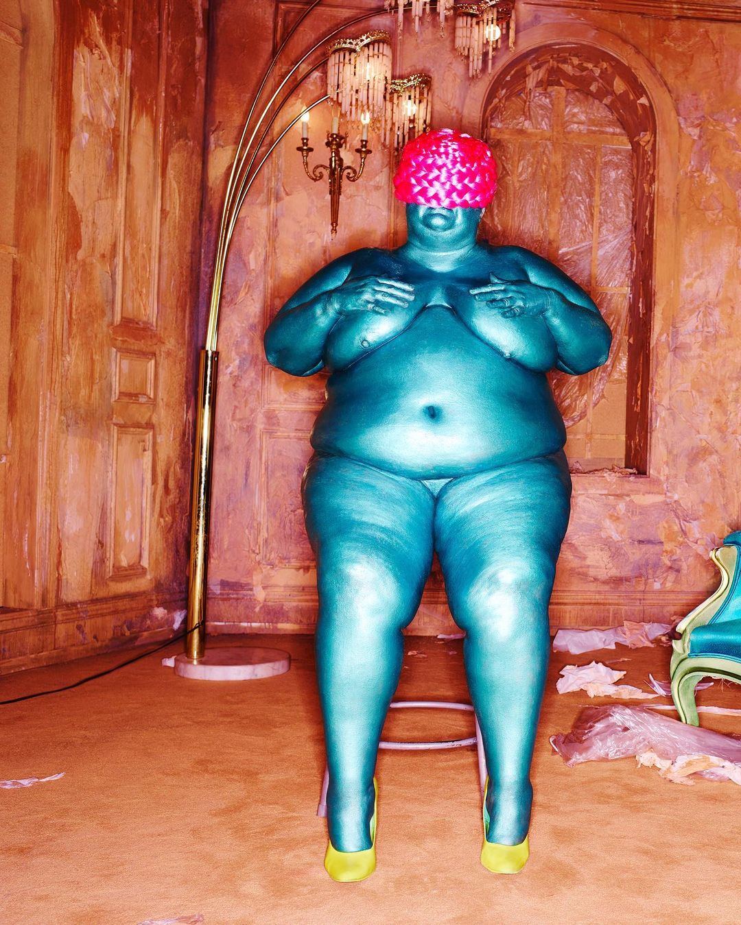 Full-body portrait of a turquoise-skinned woman wearing a pink hat, standing indoors with yellow high heels and a decorative gold lamp.