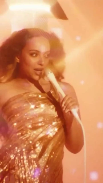 Close-up of a woman singing into a microphone, wearing a strapless gold sequin dress with an orange glow and blurred background.