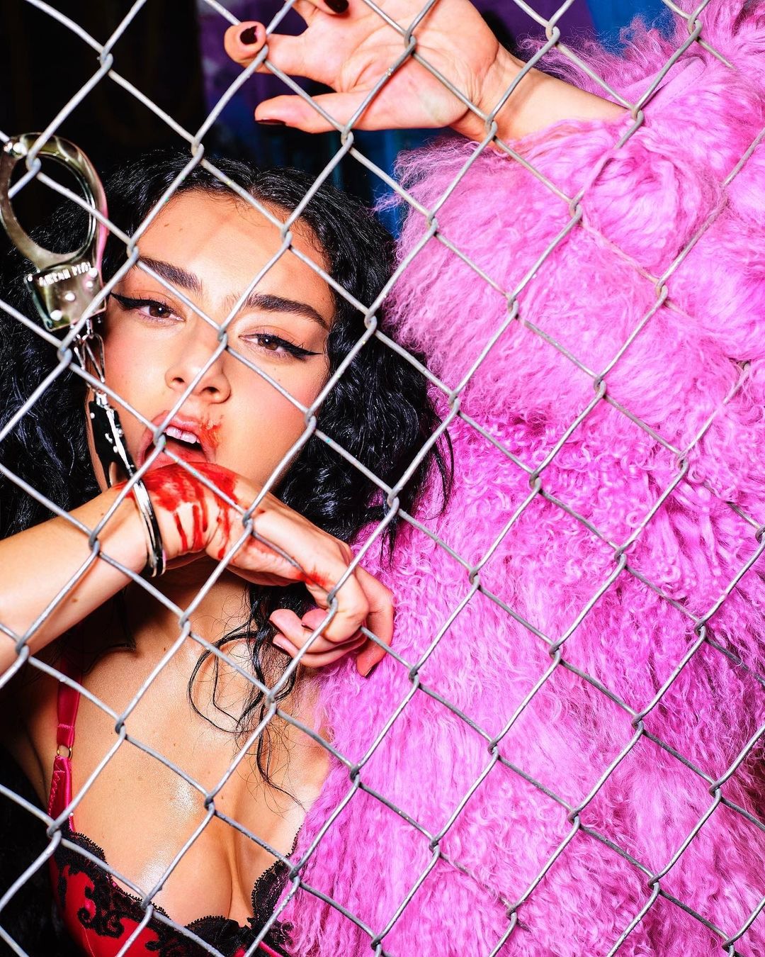 Close-up portrait of a woman in red lingerie and a fluffy pink coat behind a metal fence, with fake blood and handcuffs.