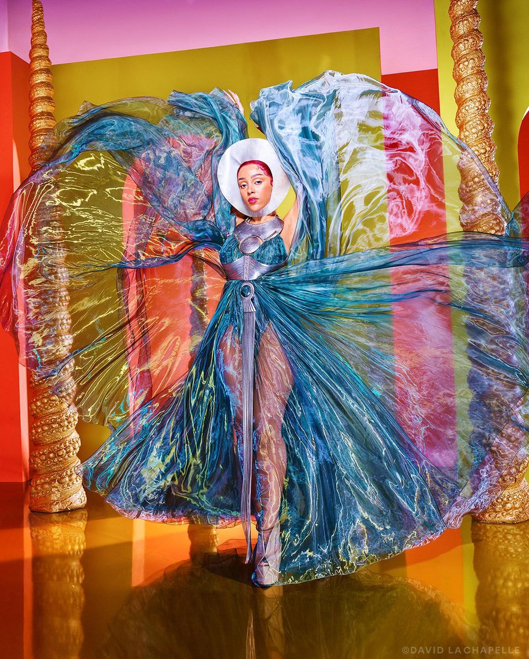 Person posing with a wide white hat and blue iridescent dress with colorful accents, posing in a glossy studio with columns.