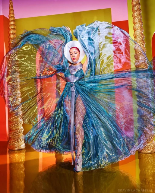 Person posing with a wide white hat and blue iridescent dress with colorful accents, posing in a glossy studio with columns.