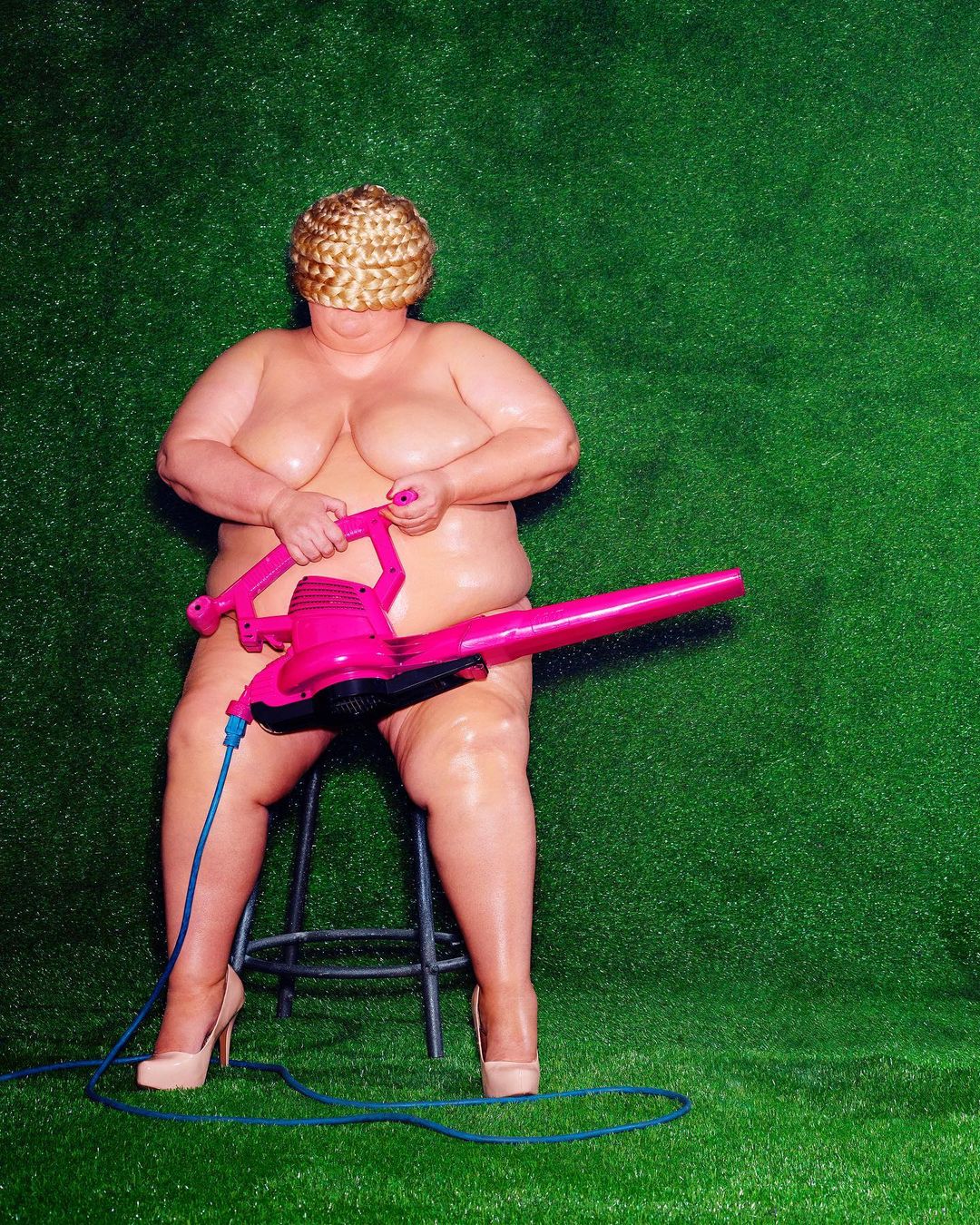 Surreal image: Overweight nude adult sits on stool holding pink leaf blower against a green background in an indoor environment.