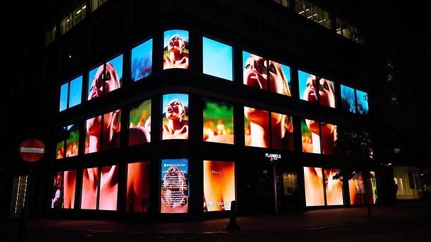 Building featuring a multi-screen video installation of a woman looking up. The storefront is illuminated and a street sign is visible.