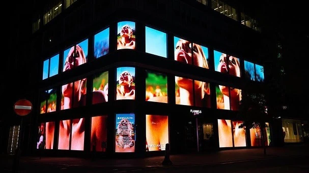 Building featuring a multi-screen video installation of a woman looking up. The storefront is illuminated and a street sign is visible.