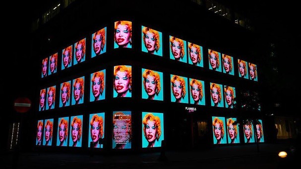 Exterior shot of a commercial building at night featuring a repeating headshot of a person with red hair on multiple screens.