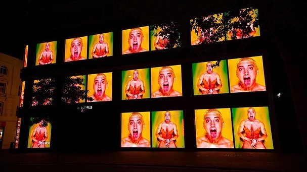 Building facade at night features a grid of large screens displaying a person with an open mouth and painted skin. Trees visible on building.