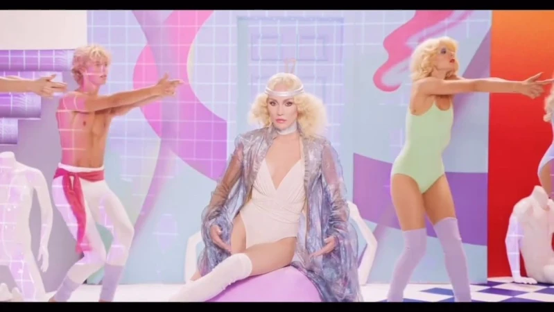 A woman with blonde hair wearing a shiny jacket seated on a ball, surrounded by dancers in a colorful and retro studio setting.