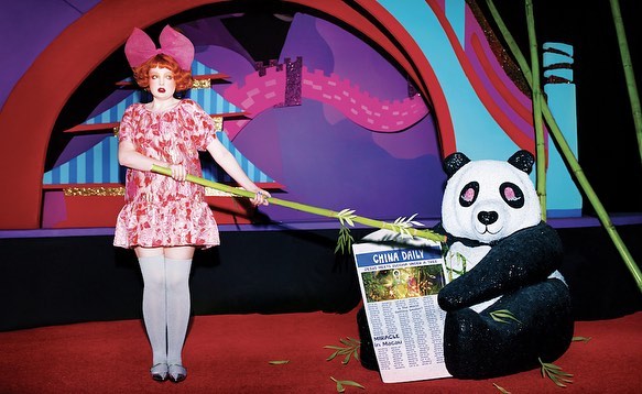 Adult female in a pink floral dress holds bamboo next to a panda bear reading a newspaper in a whimsical studio environment.