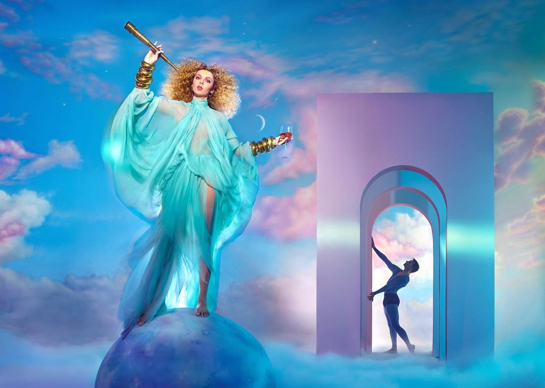 Woman in flowing turquoise dress with telescope stands on a globe with a man in doorway against a cloudy sky backdrop.