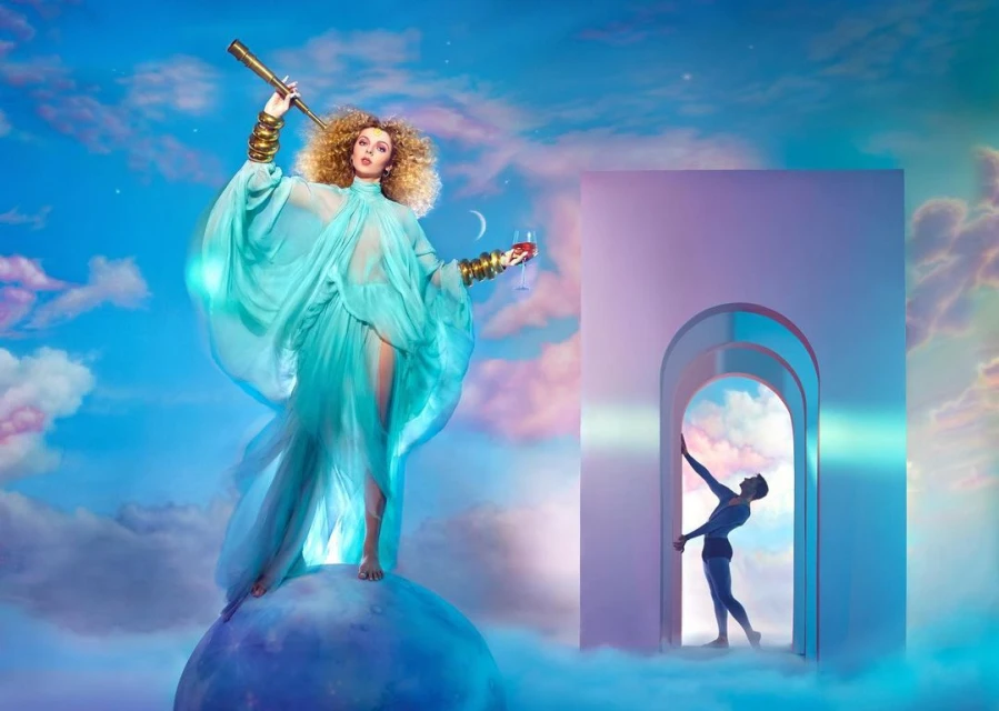 Woman in flowing turquoise dress with telescope stands on a globe with a man in doorway against a cloudy sky backdrop.