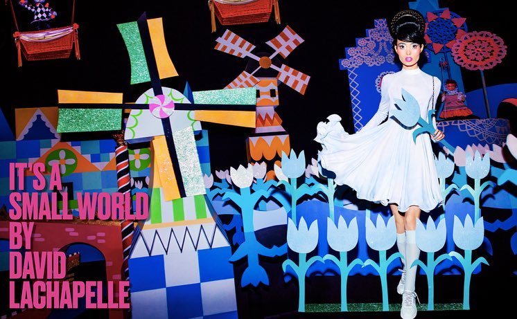 Fashion shot of a woman in a white dress, posing in front of colorful, graphic, cartoon-like set pieces with geometric shapes and windmill.