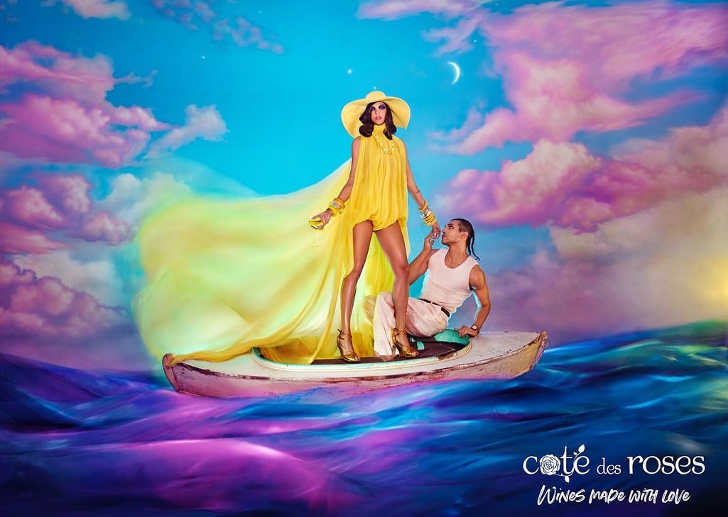 An adult woman in a yellow dress stands in a boat with a man, with a vivid sky and water background in an advertisement.