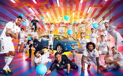 Group of male athletes in white uniforms posing with a blue electric car in a surreal pink and yellow striped room