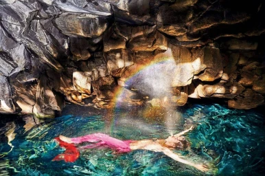 Person floating in vibrant turquoise water inside a rocky grotto with a natural prism rainbow arc shimmering above them.