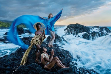 Three figures in theatrical costumes posed on dark volcanic rocks against a crashing ocean and stormy sky, photographed by David Lachapelle.