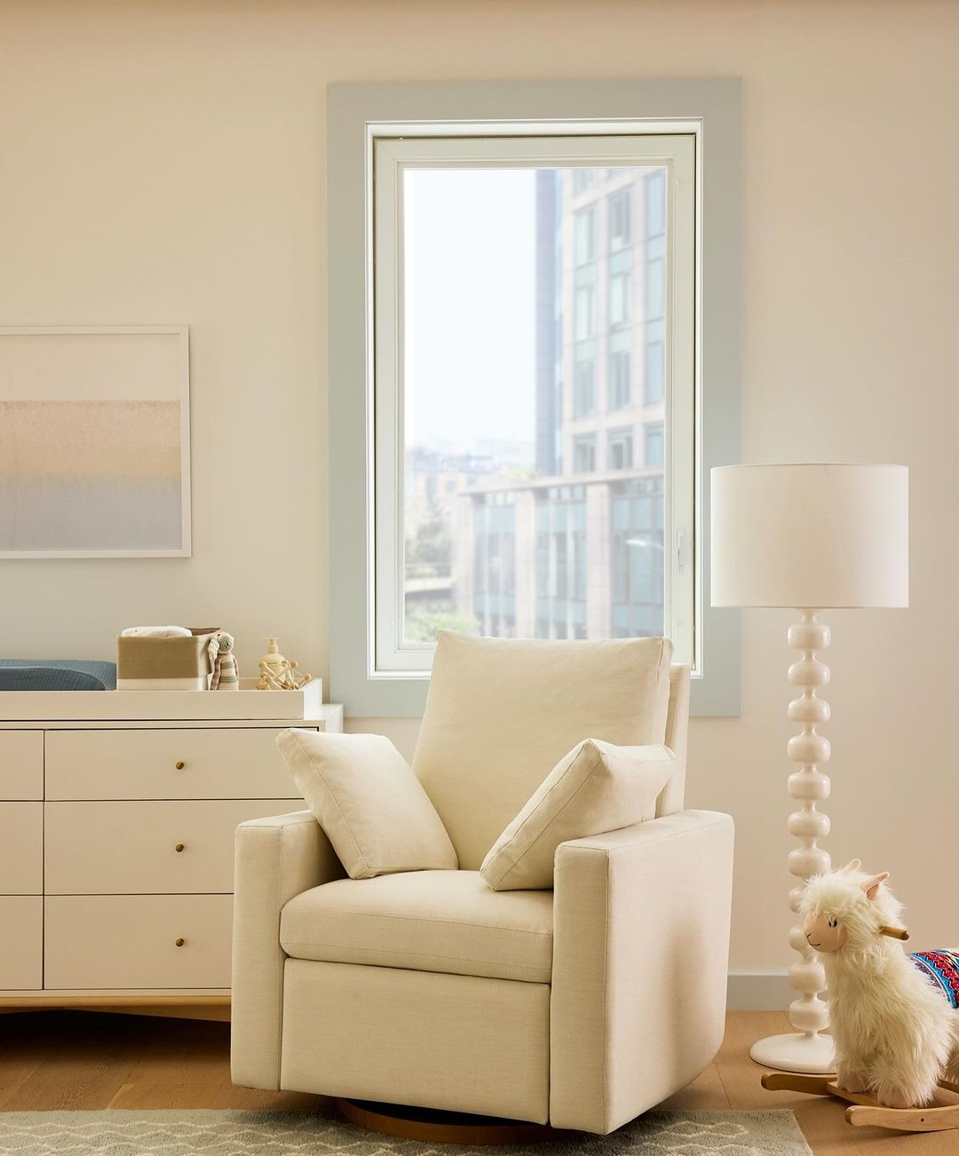 Interior shot of a nursery with a modern armchair, light wood dresser, sheep rocking horse toy, and large window with city view.