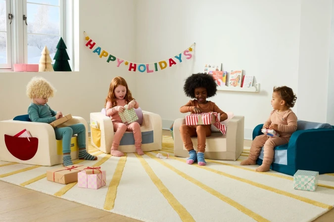 Four young children are seated in small chairs opening gifts in a brightly lit room with a colorful rug and a holiday banner.