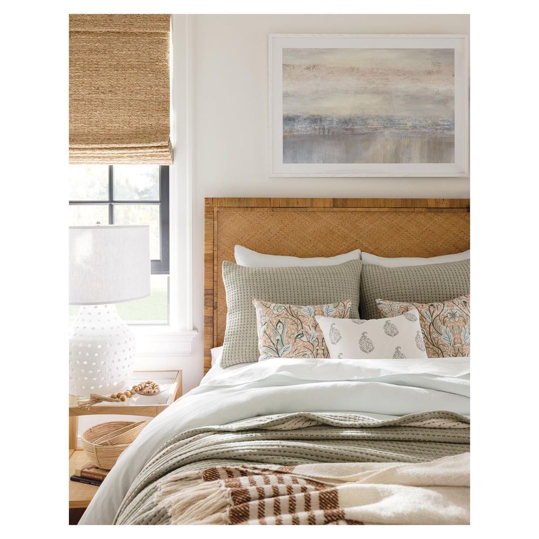 Bedroom features a bed with woven headboard, pillows, throw blanket, and nightstand with lamp. Natural light fills the neutral space.
