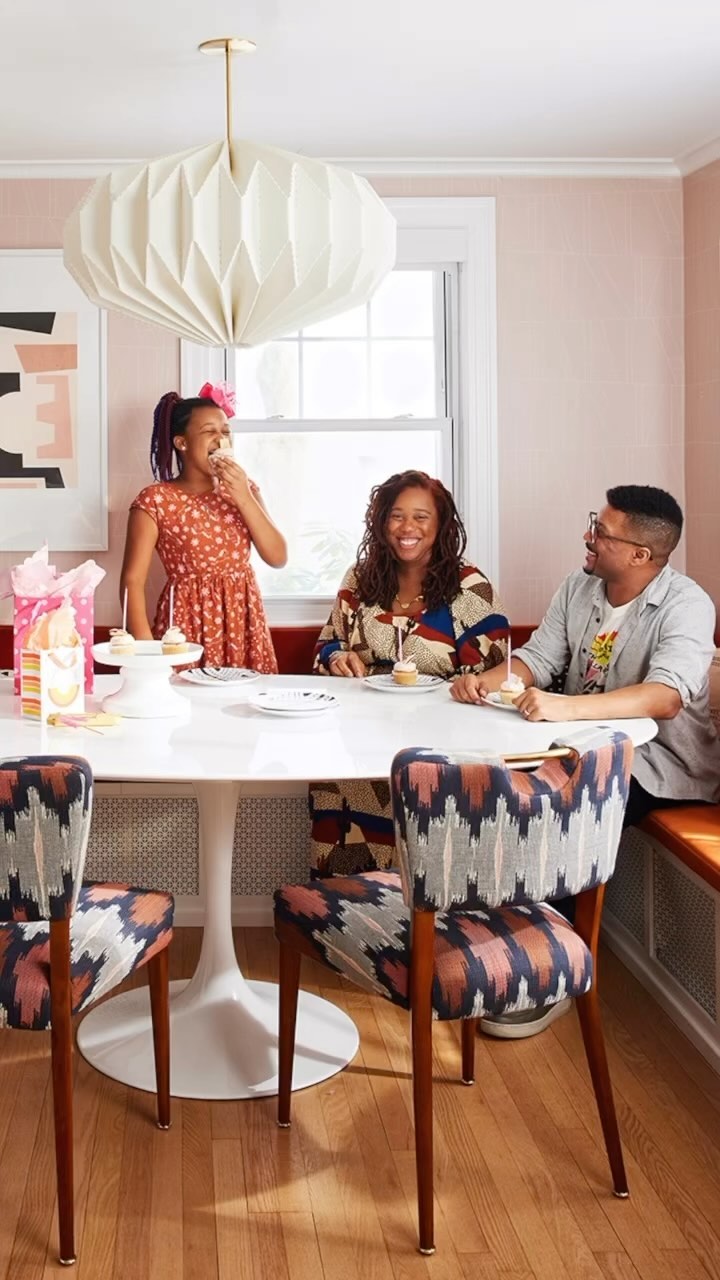 Three people—adult female, adult male, and child—sit around a white table with cupcakes in an interior with pink walls and wood floors.