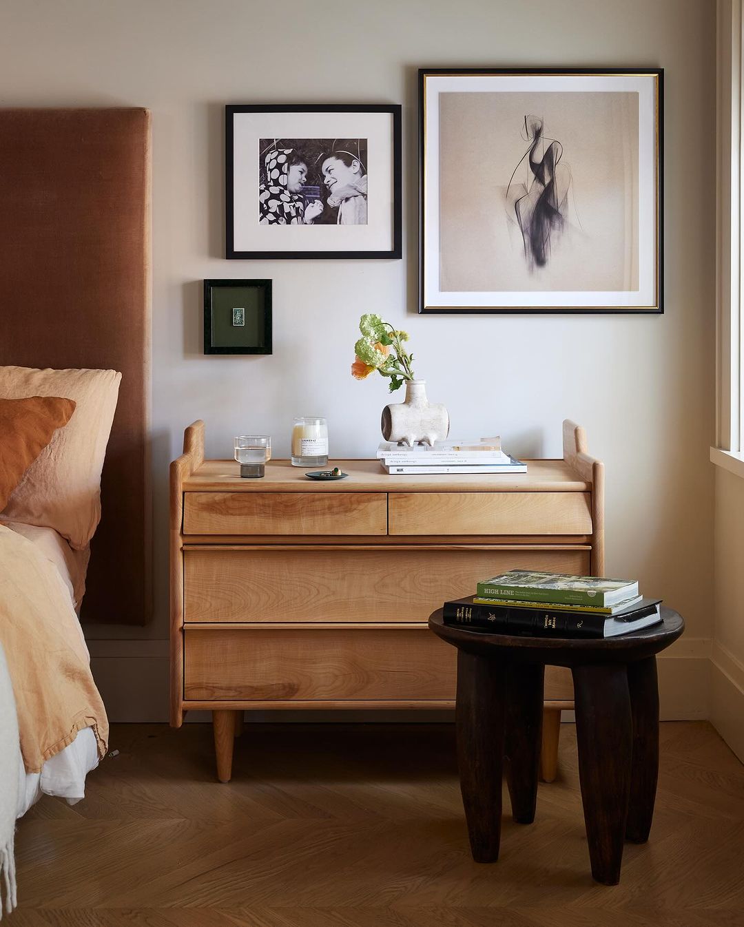 Bedroom interior with wooden nightstand, artwork, books, bedside lamp, and flowers. Stylish design with warm colors and natural light.