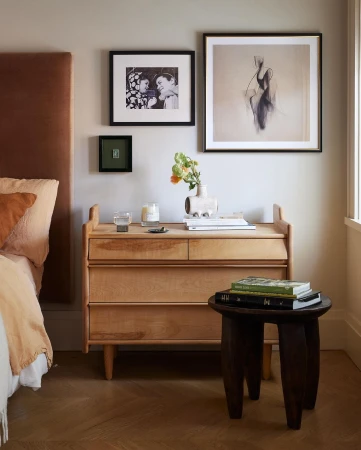 Bedroom interior with wooden nightstand, artwork, books, bedside lamp, and flowers. Stylish design with warm colors and natural light.