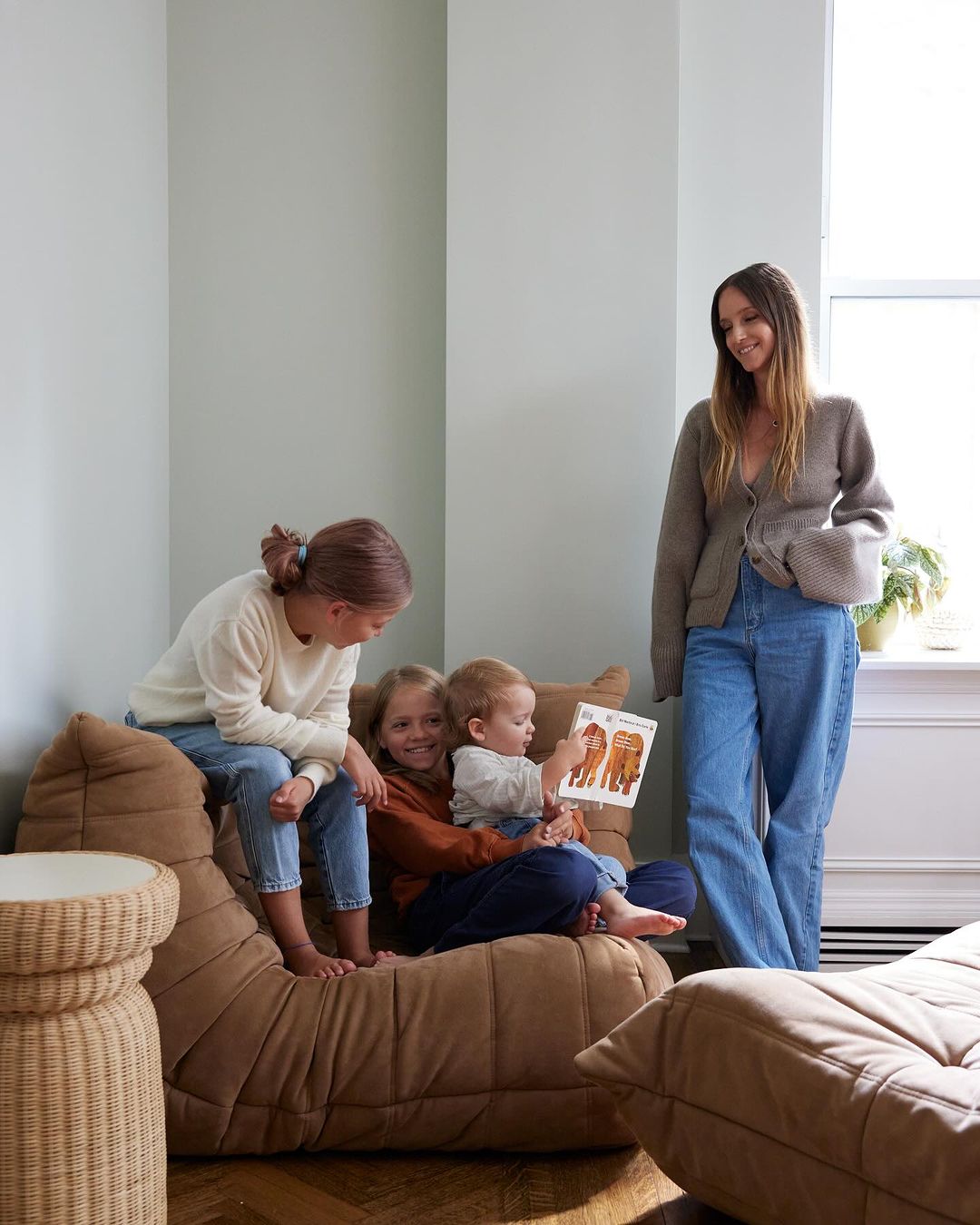 A mother sits with three kids on a brown sofa in a naturally lit room; she reads from a book with animal illustrations.