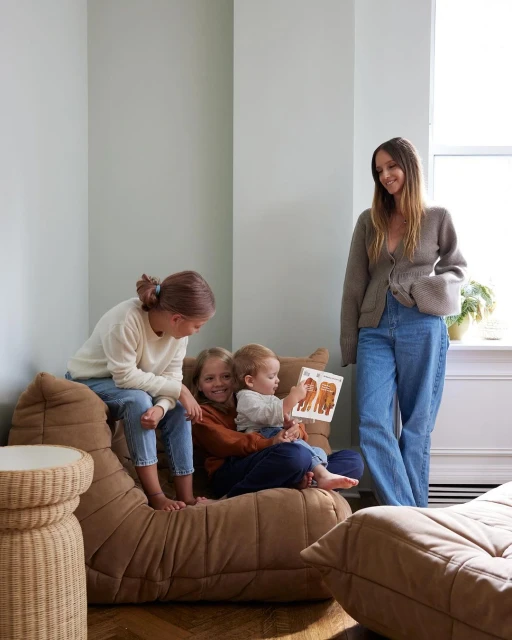 A mother sits with three kids on a brown sofa in a naturally lit room; she reads from a book with animal illustrations.