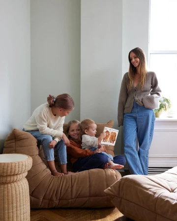 A mother sits with three kids on a brown sofa in a naturally lit room; she reads from a book with animal illustrations.