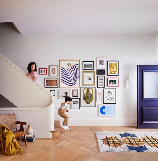 A boy jumps down stairs in a modern home with a woman looking on from above, surrounded by framed art on a white wall.