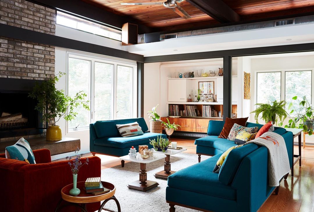Bright and spacious living room with teal sofas, orange armchairs, a brick fireplace, and large windows for natural lighting.