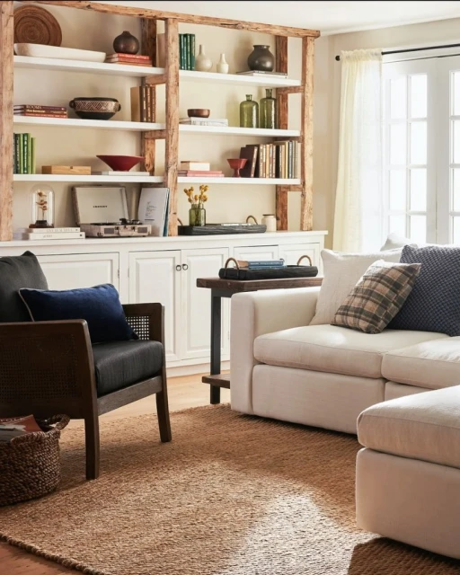 Living room interior with a white couch, dark accent chair, and a textured brown rug. A bookshelf displays various decorative objects.