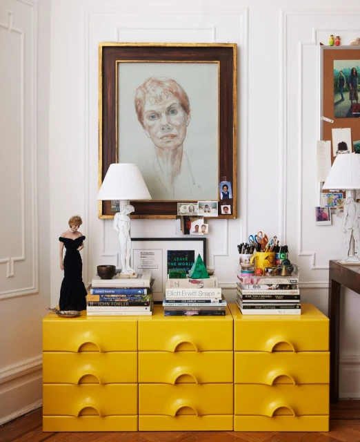 Interior view featuring a yellow dresser adorned with books, lamps, artwork, and decorative items, set against a white wall.