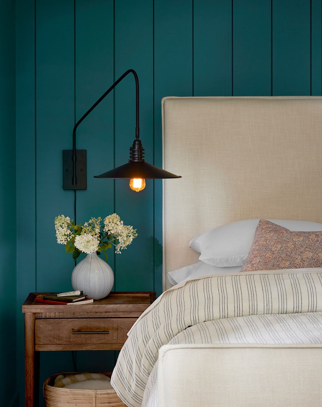 Bedroom interior with a bed, wooden nightstand, and a wall-mounted lamp against a teal wall, creating a cozy and inviting space.