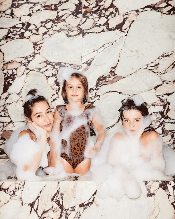 Woman and two children enjoy bath time in a marble-clad bathroom filled with bubbly soap, creating a moment of family fun and relaxation.