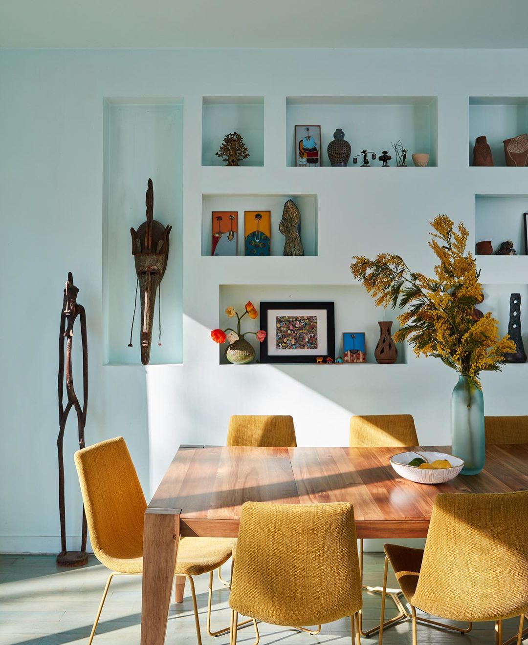 Interior of a dining room with a wood table and yellow chairs, featuring art and decor items displayed in recessed wall niches.