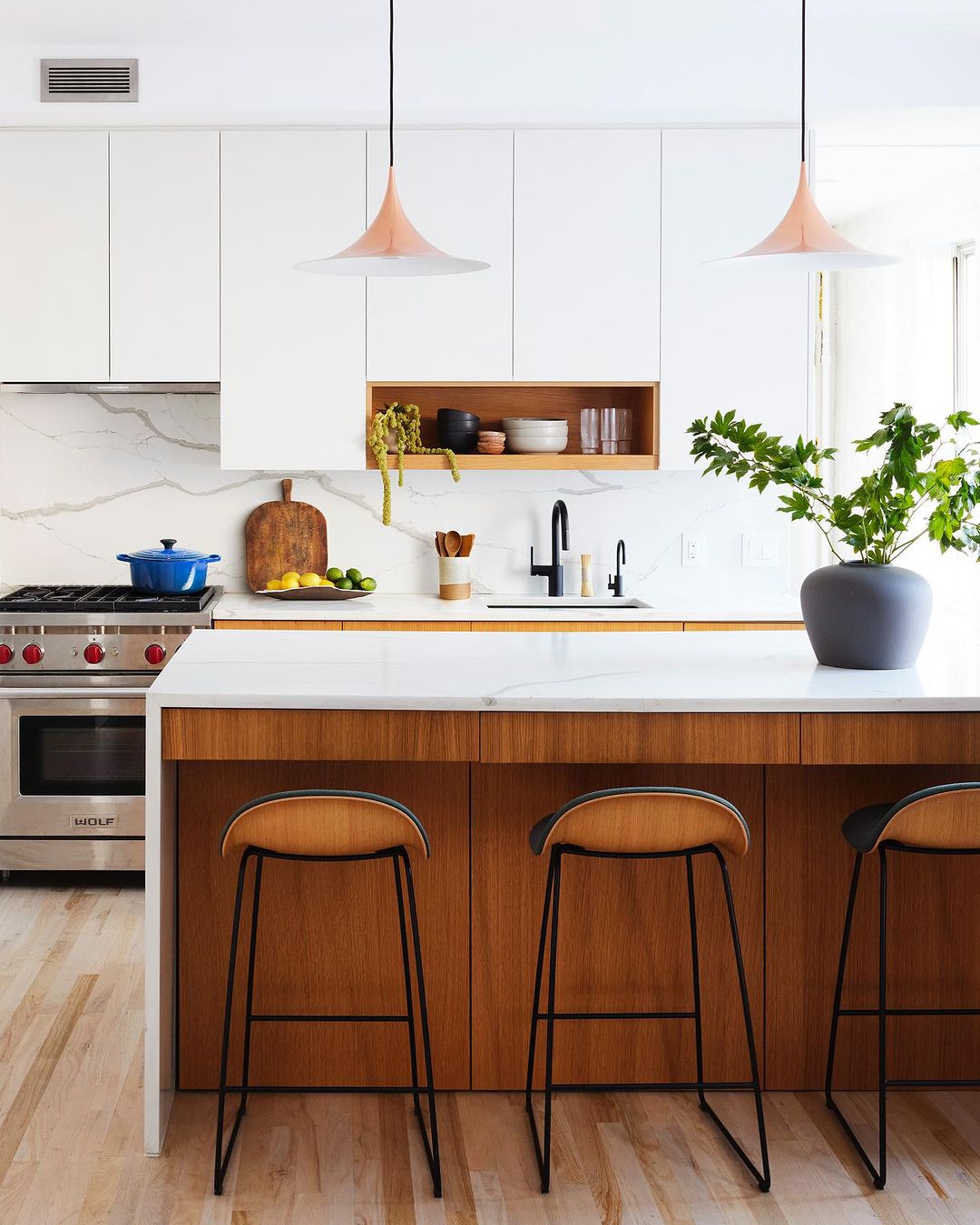 Modern kitchen interior with white cabinets, marble countertops, wood island with stools, pendant lights, and stainless steel appliances.
