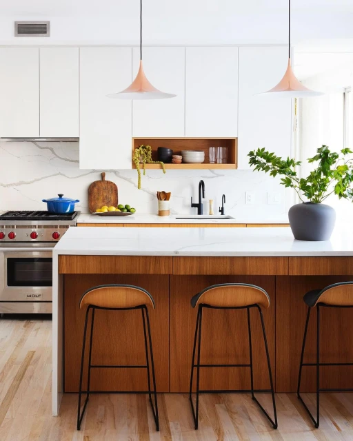 Modern kitchen interior with white cabinets, marble countertops, wood island with stools, pendant lights, and stainless steel appliances.