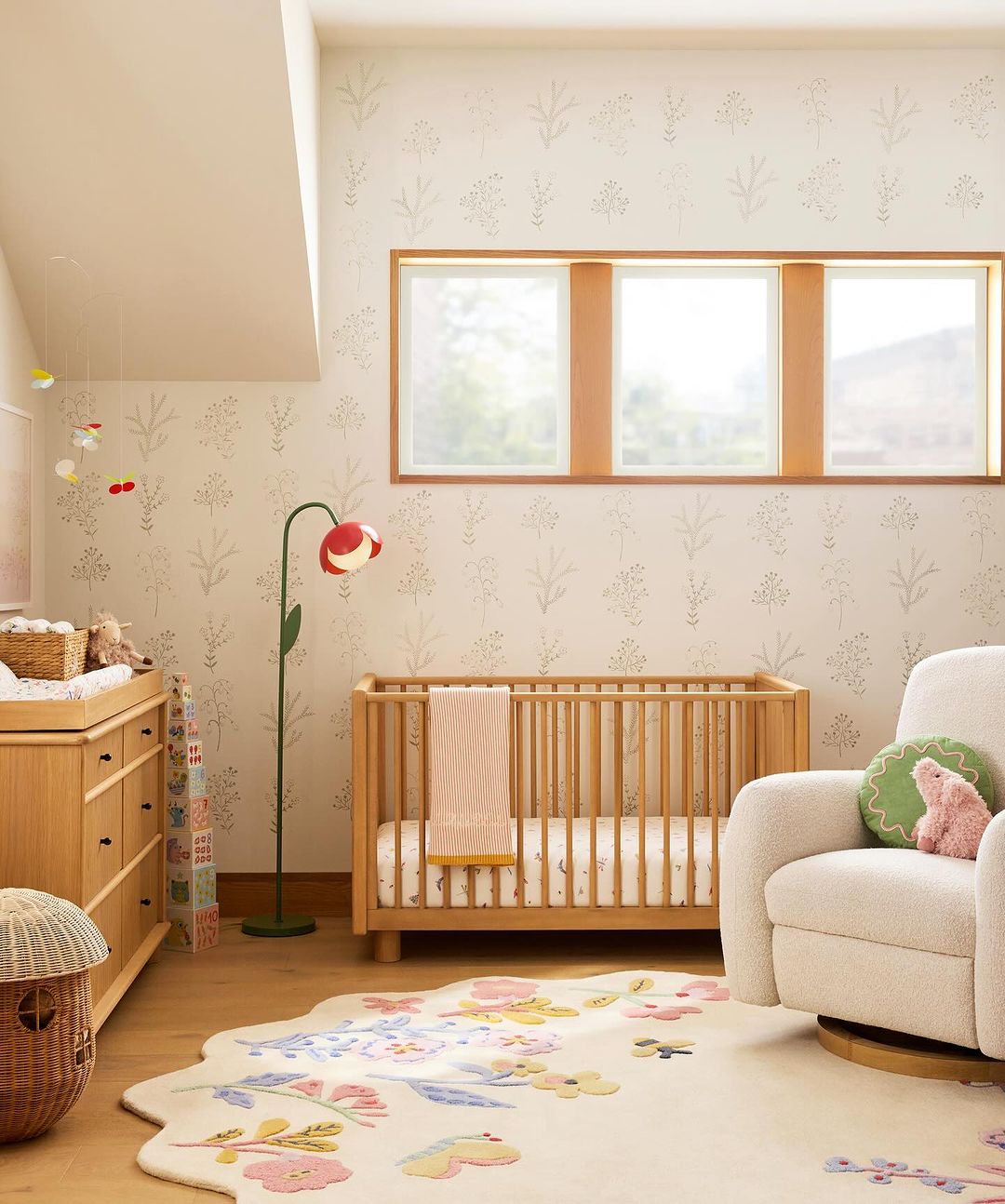 Nursery interior with a light wood crib, changing table, and floral rug, illuminated by natural light from windows.