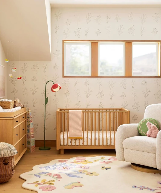Nursery interior with a light wood crib, changing table, and floral rug, illuminated by natural light from windows.