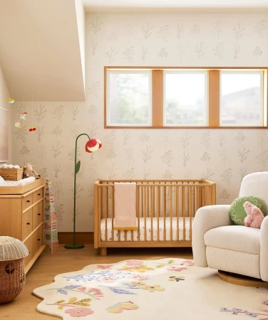 Nursery interior with a light wood crib, changing table, and floral rug, illuminated by natural light from windows.