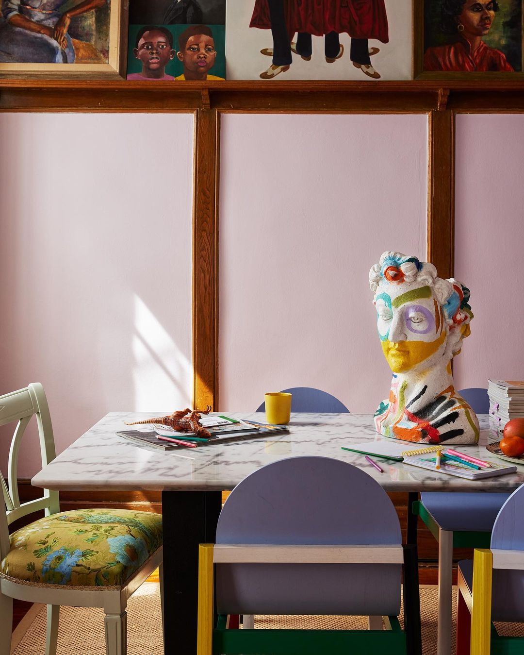 Interior with a marble table surrounded by colorful chairs, adorned with a bust sculpture and paintings hanging on the wall.