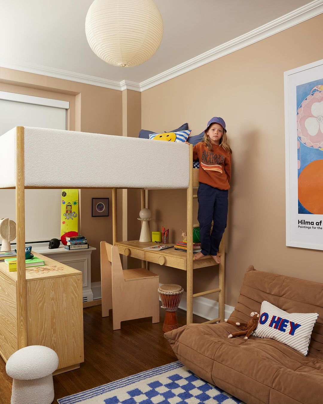Child stands on the ladder of a loft bed in a bedroom with hardwood floors, a beige futon, a wooden desk, and a blue checkered rug.