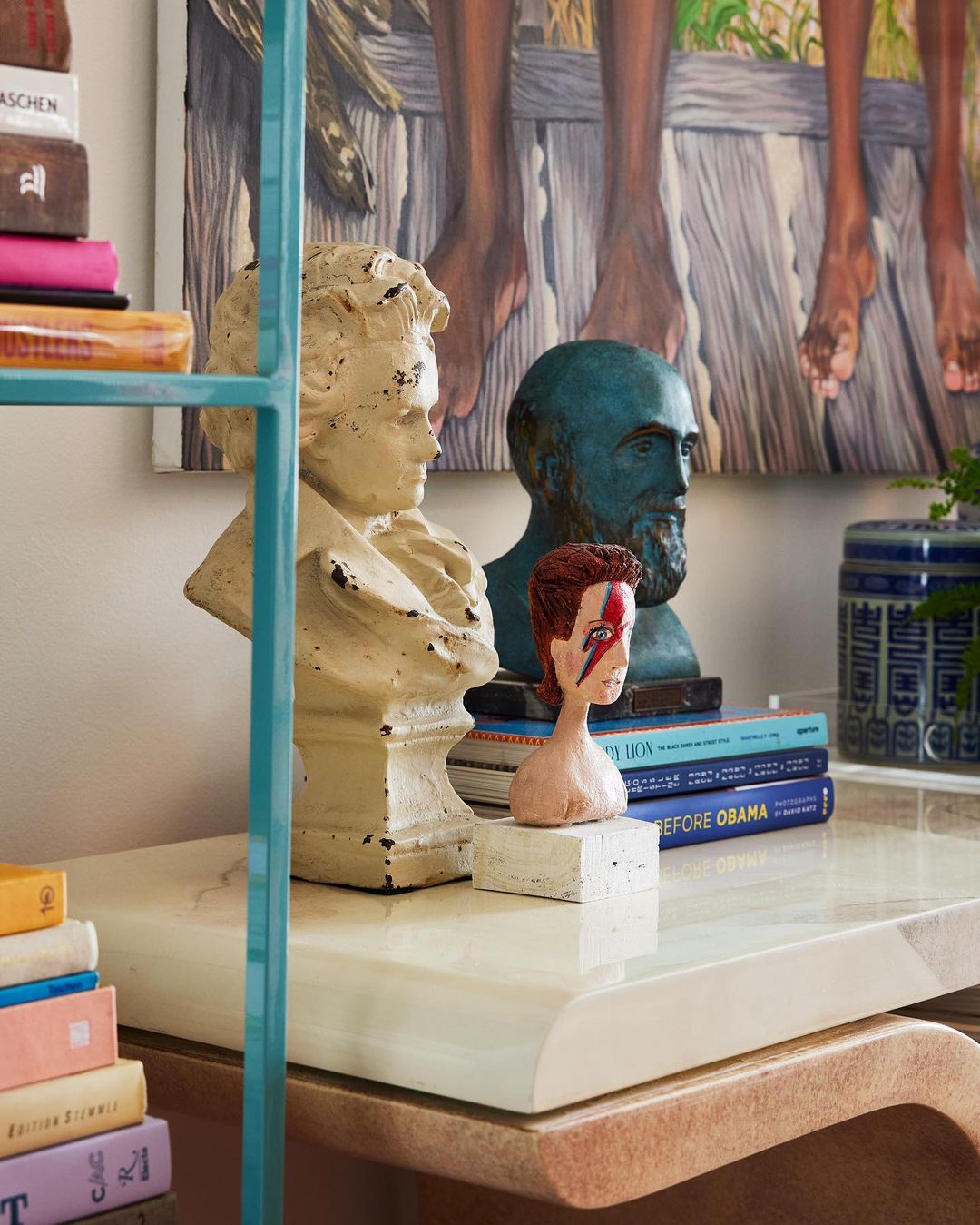 Interior design featuring a marble-topped table, an open bookshelf, decorative busts, books, and a painting of people's legs.