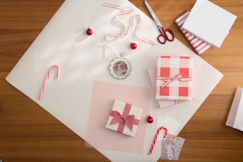 Overhead shot of Christmas gifts being wrapped on a wooden table with ornaments, ribbon, scissors, and candy canes, gift wrapping paper.