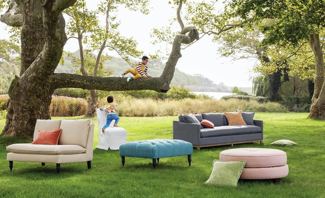 Two children on tree, one climbing chair; sofas, cushions arranged on lawn near trees, with body of water in background on sunny day.