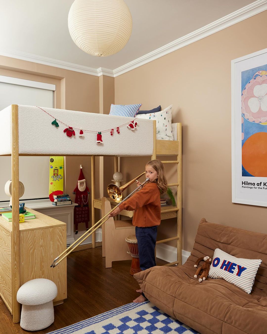 Young child plays a brass trombone in a warmly-lit bedroom with an elevated loft bed, desk, soft brown sofa, and blue checkered rug.