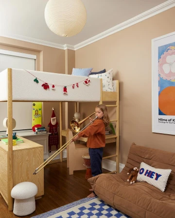 Young child plays a brass trombone in a warmly-lit bedroom with an elevated loft bed, desk, soft brown sofa, and blue checkered rug.