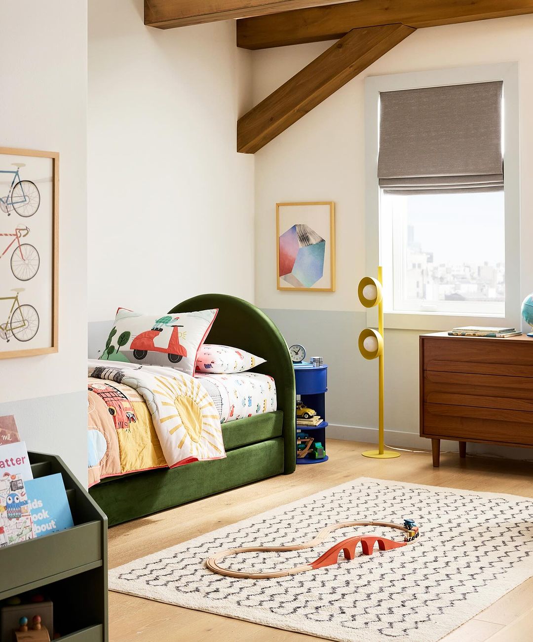 Olive green platform bed with colorful quilt and throw pillows in child's room with wooden beams, window, neutral colors, and hardwood floors.
